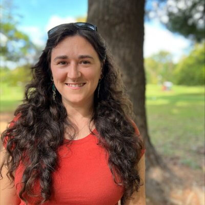 The image shows a woman with long, curly brown hair, wearing a red t-shirt. She is smiling and standing outside, with a tree trunk visible behind her. The background features a green lawn and some trees, suggesting a park or garden setting. She has sunglasses on top of her head.